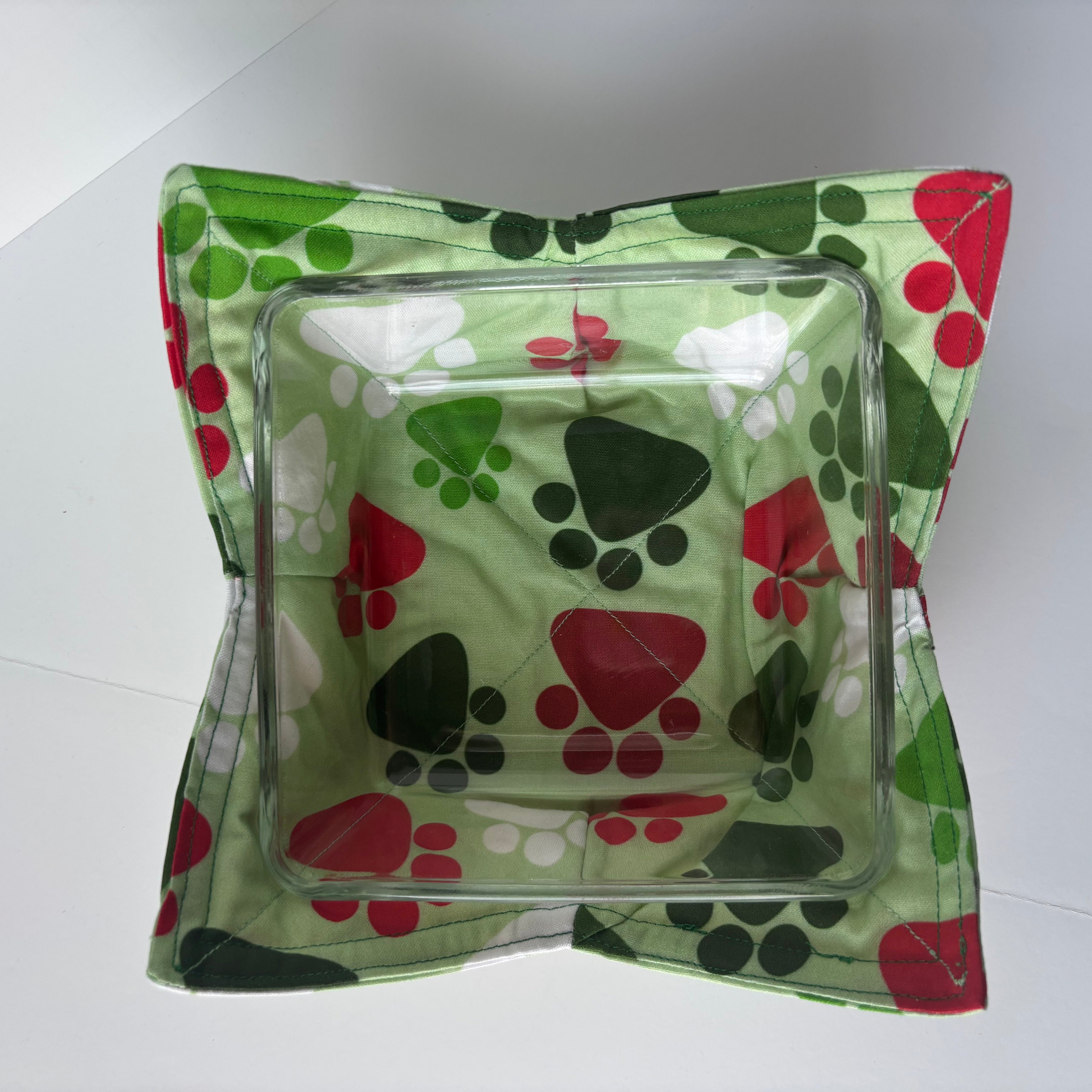 Clear glass square bowl with a green fabric cozy bowlfeaturing paw prints on a white background