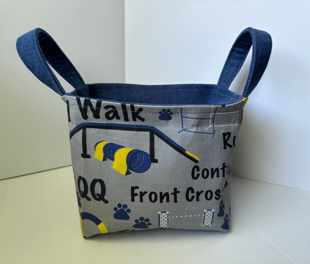 A fabric storage basket with two blue handles, featuring a gray printed exterior with dog exercise-themed text and graphics, and a blue fabric lining.