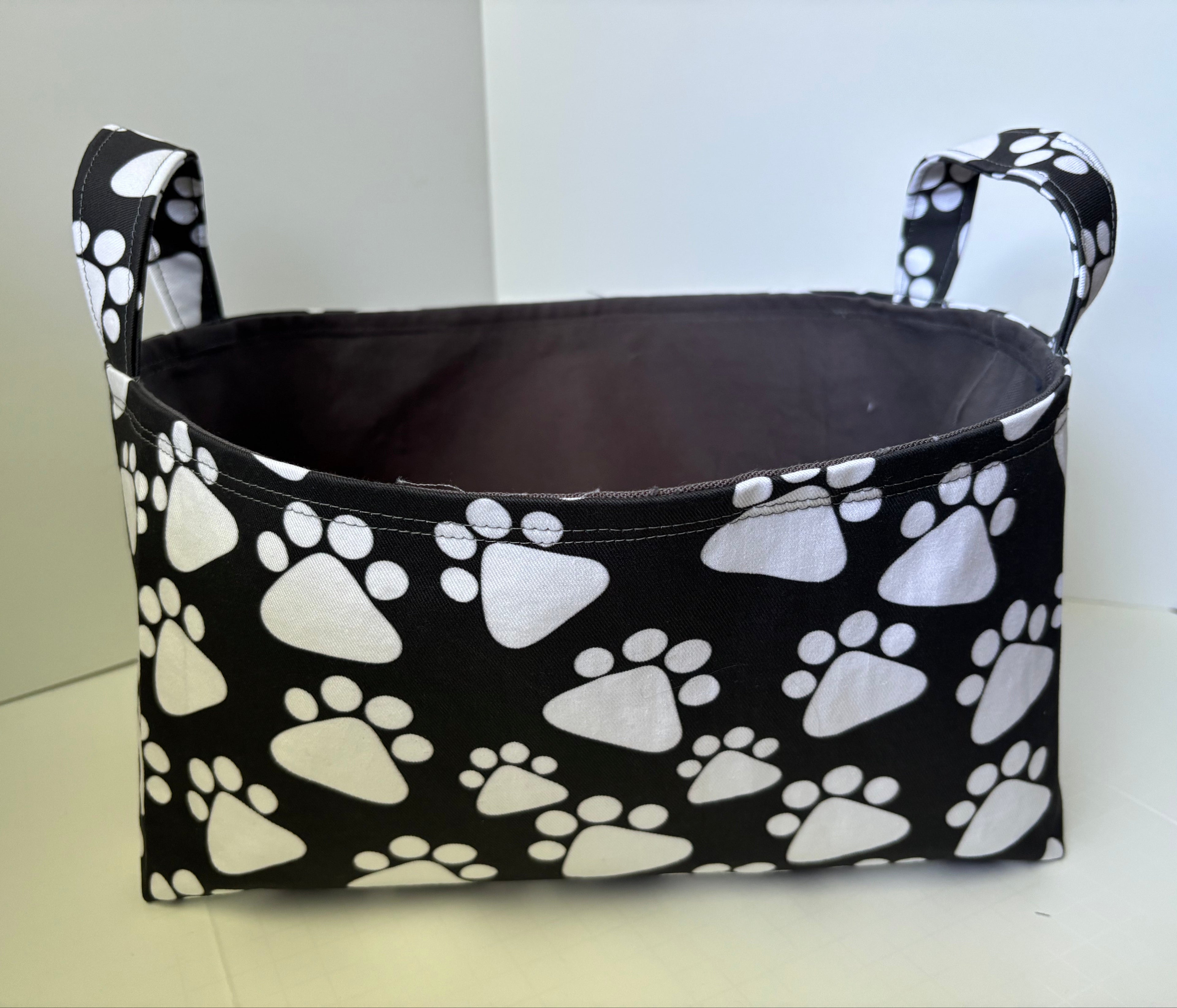 A fabric storage basket with a black background and white paw print pattern, featuring two top handles and a dark gray interior lining, set against a plain light background.