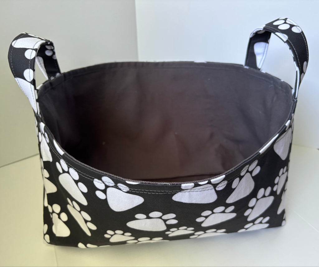 A fabric storage basket with a black exterior printed with white paw prints and a dark gray interior, shown on a light-colored surface.
