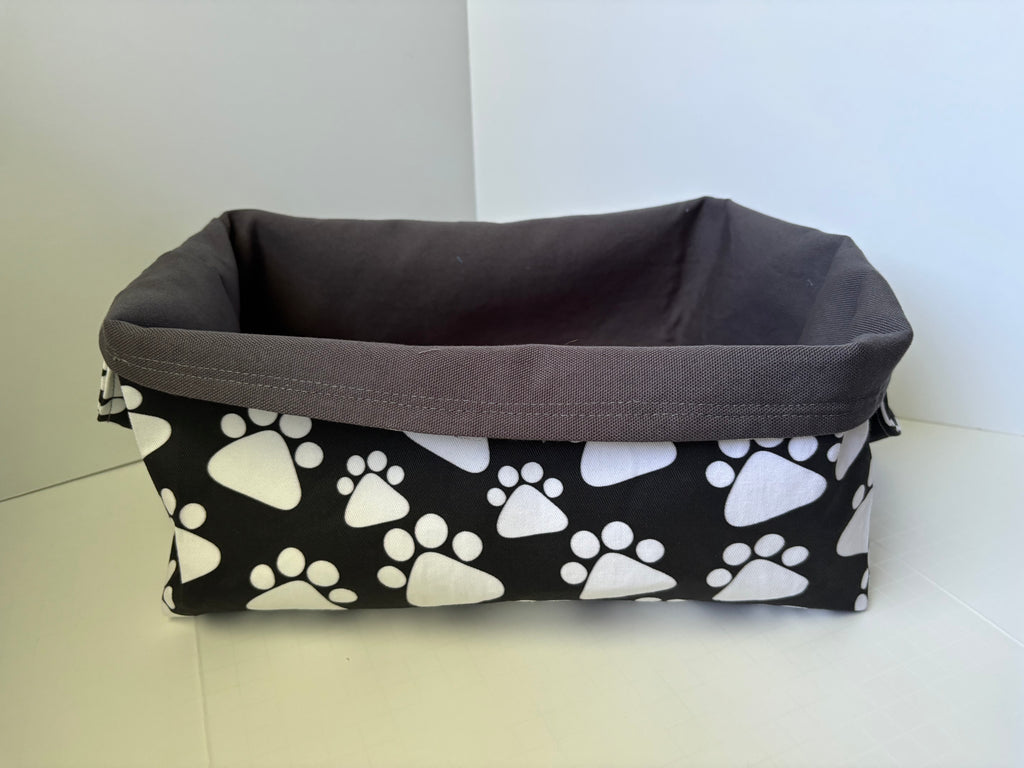 A rectangular fabric storage basket with a black and white paw print pattern on the exterior and a solid dark gray fabric interior, shown on a light-colored floor against a white wall.