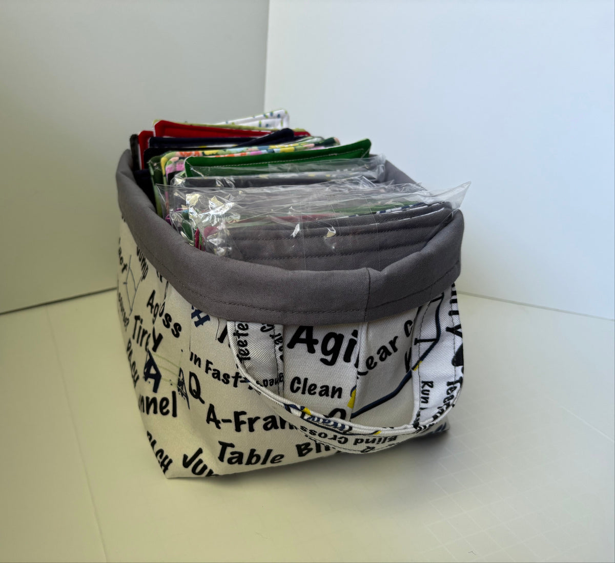 A fabric storage basket with a gray fabric lining and a white printed fabric exterior featuring black text and diagrams. The basket is filled with various sewing or crafting supplies, including a clear plastic bag containing green and red fabric pieces.