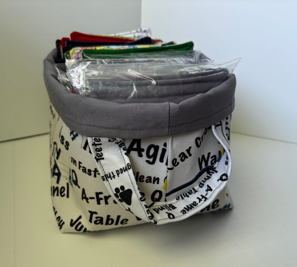 A fabric storage basket with a gray fabric top and a white printed fabric body featuring black text and paw prints. The basket is filled with various items, including a clear plastic bag of small items, and sits on a light-colored surface against a plain background.