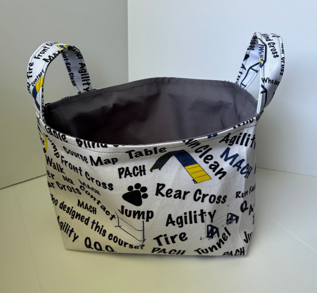 A fabric storage basket with dual handles, featuring a white printed exterior with black text and graphics related to dog agility, and a dark gray interior.
