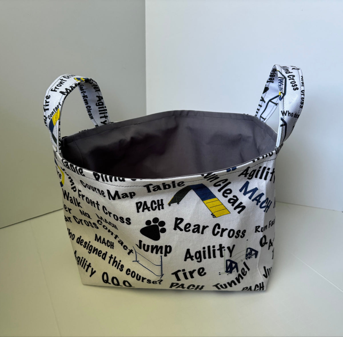 A fabric storage basket with dual handles, featuring a white printed exterior with black text and graphics related to cross country running, and a dark gray interior.