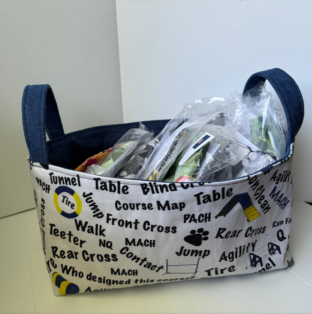 A fabric storage basket with dark blue handles, filled with packaged items. The basket features a white printed pattern with black text and logos related to motocross or racing, including 'Tire', 'Jump', 'Front Cross', 'Rear Cross', and 'Agility'.
