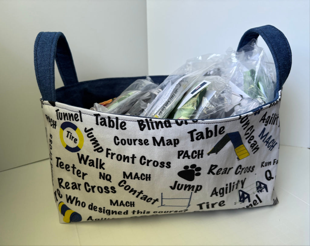 A fabric storage basket with blue denim handles and a cream canvas body printed with black dog agility course terms like 'Table', 'Cross', and 'Jump'. The basket is filled with packaged items and photographed against a plain off-white background.