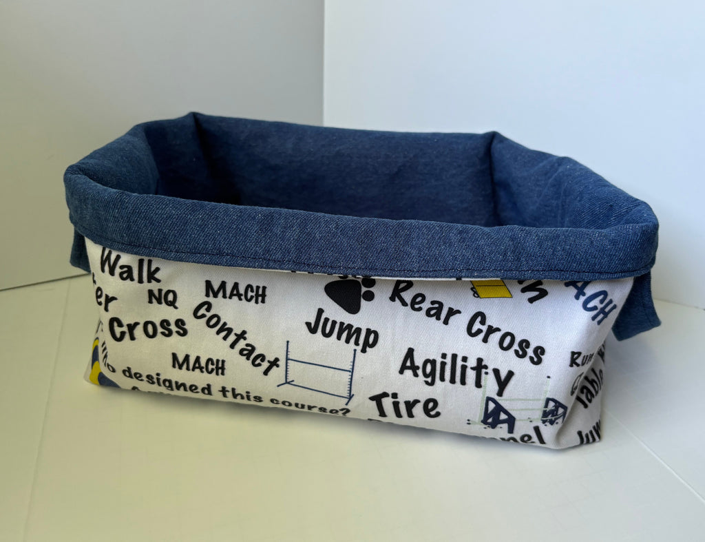 A rectangular fabric pet basket with a blue denim-like interior and exterior trim, and a white printed exterior panel featuring black racing and cross terms such as 'Mach', 'Jump', 'Rear Cross', and 'Tire'.
