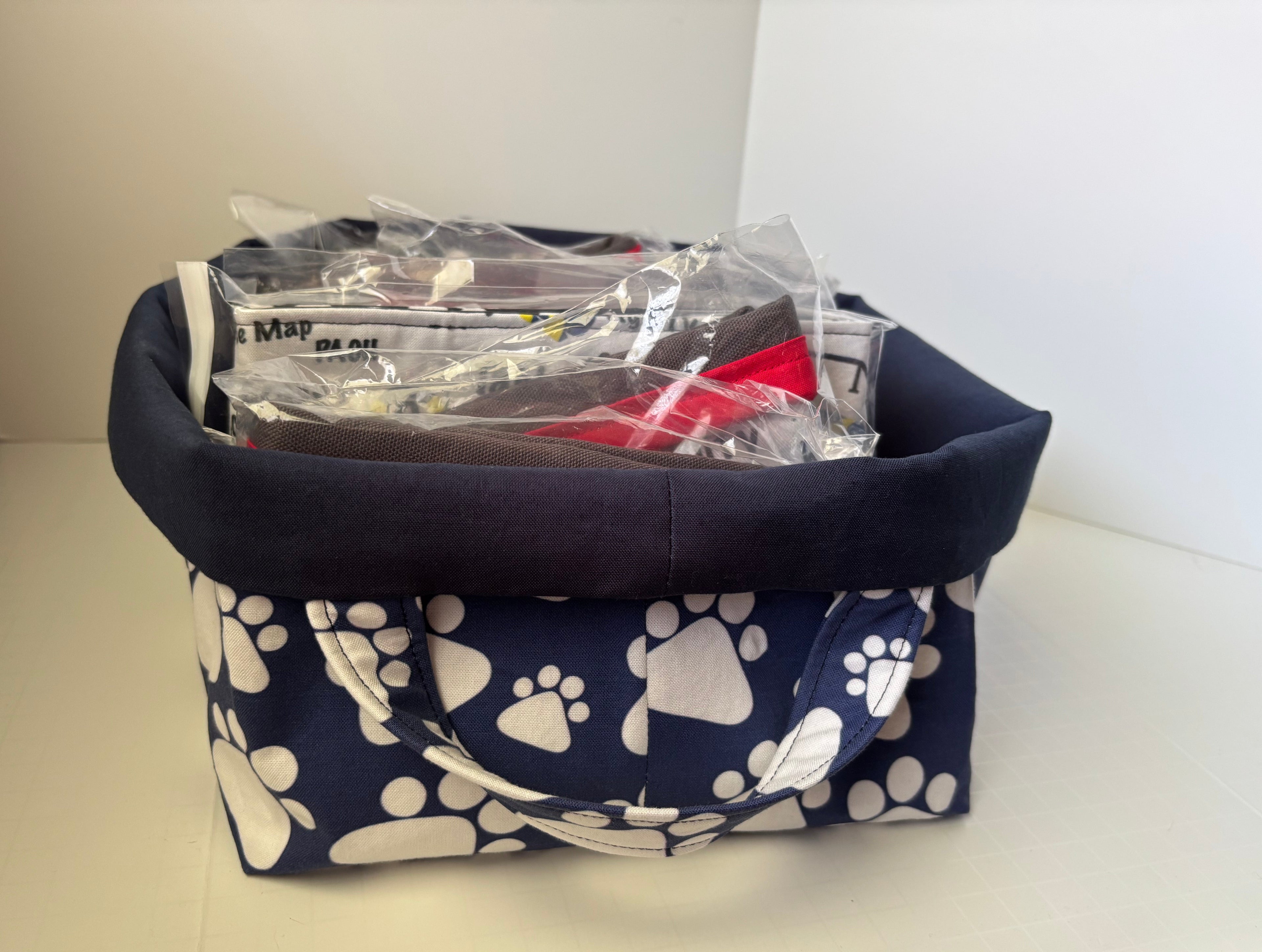 A fabric storage basket with a navy blue base printed with white paw prints, featuring a solid navy blue top rim and handles. The basket is shown filled with various packaged items on a white surface against a white wall.