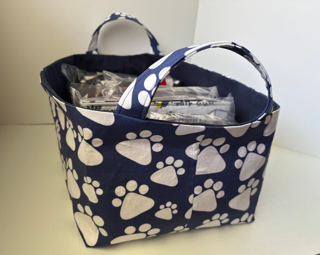 A fabric storage basket with a navy blue and white paw print pattern, featuring two top handles and filled with packaged items, set on a light-colored surface.