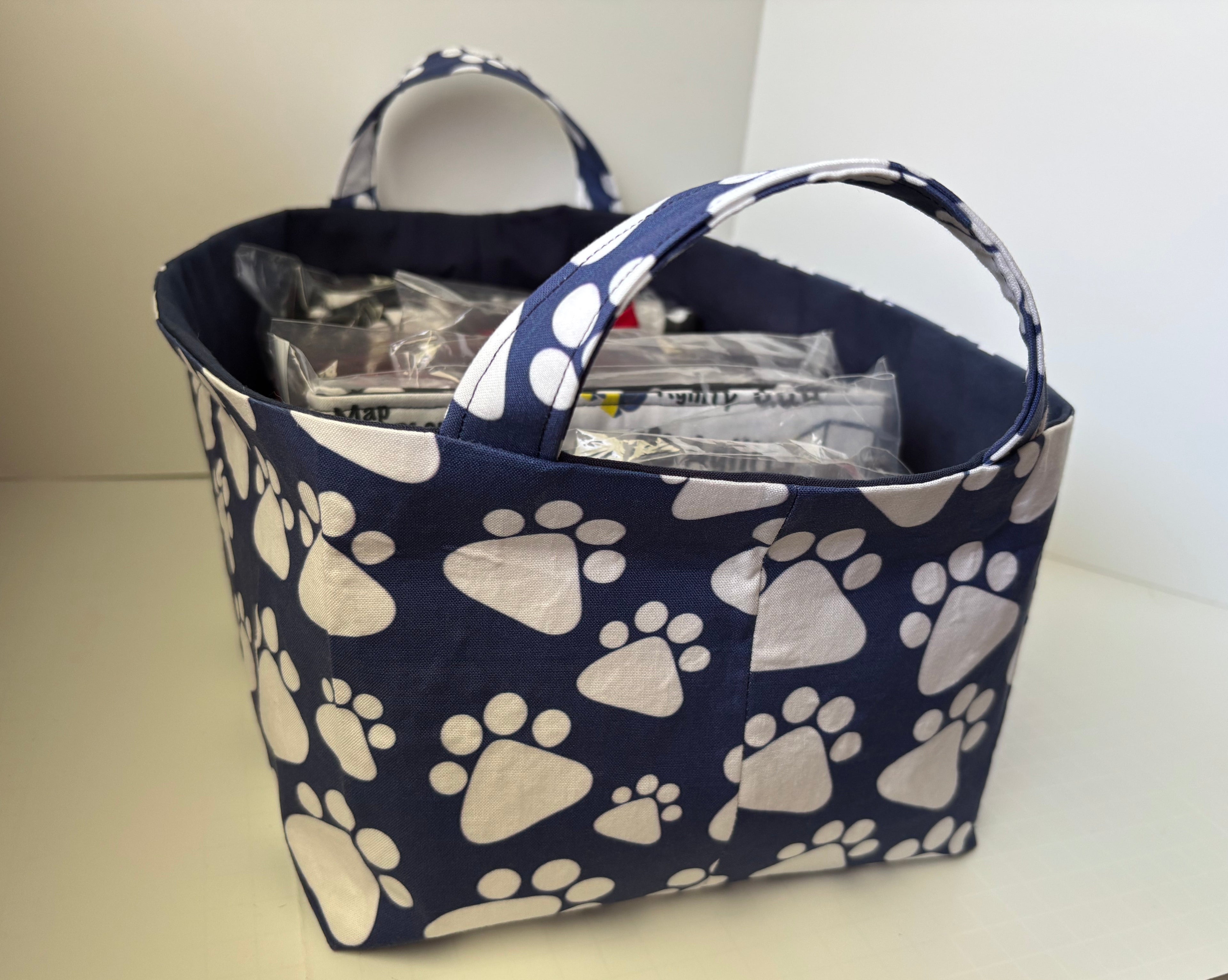 A fabric storage basket with a navy blue and white paw print pattern, featuring two top handles and filled with packaged items, set on a light-colored surface.