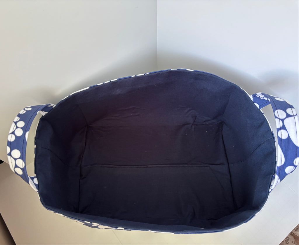 A fabric storage basket viewed from above, featuring a dark navy blue interior and blue-and-white paw print patterned fabric handles, set against a plain light gray background.