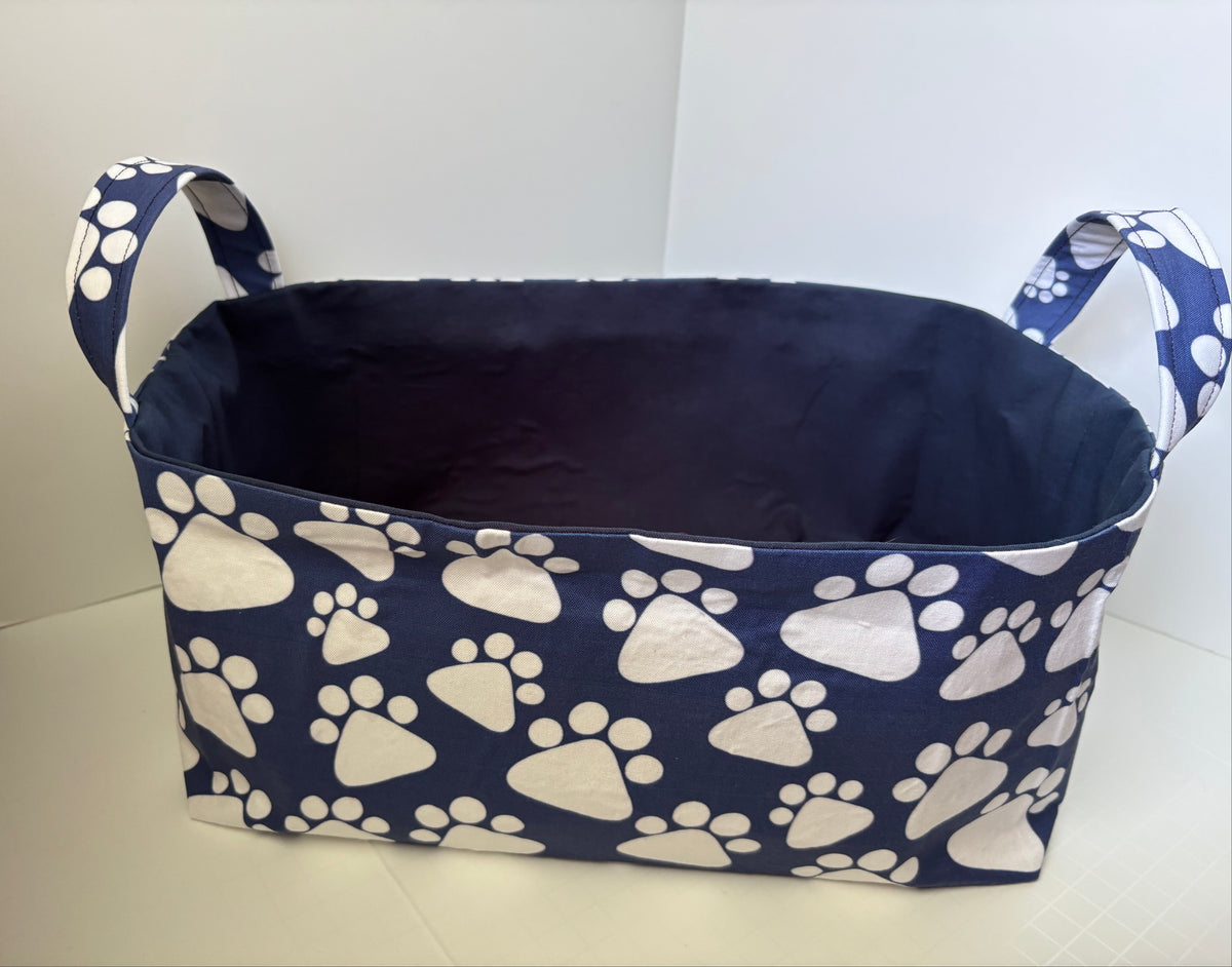 A fabric storage basket with a navy blue base printed with white paw prints, featuring two side handles and a dark navy interior, shown on a light-colored floor against a white wall.