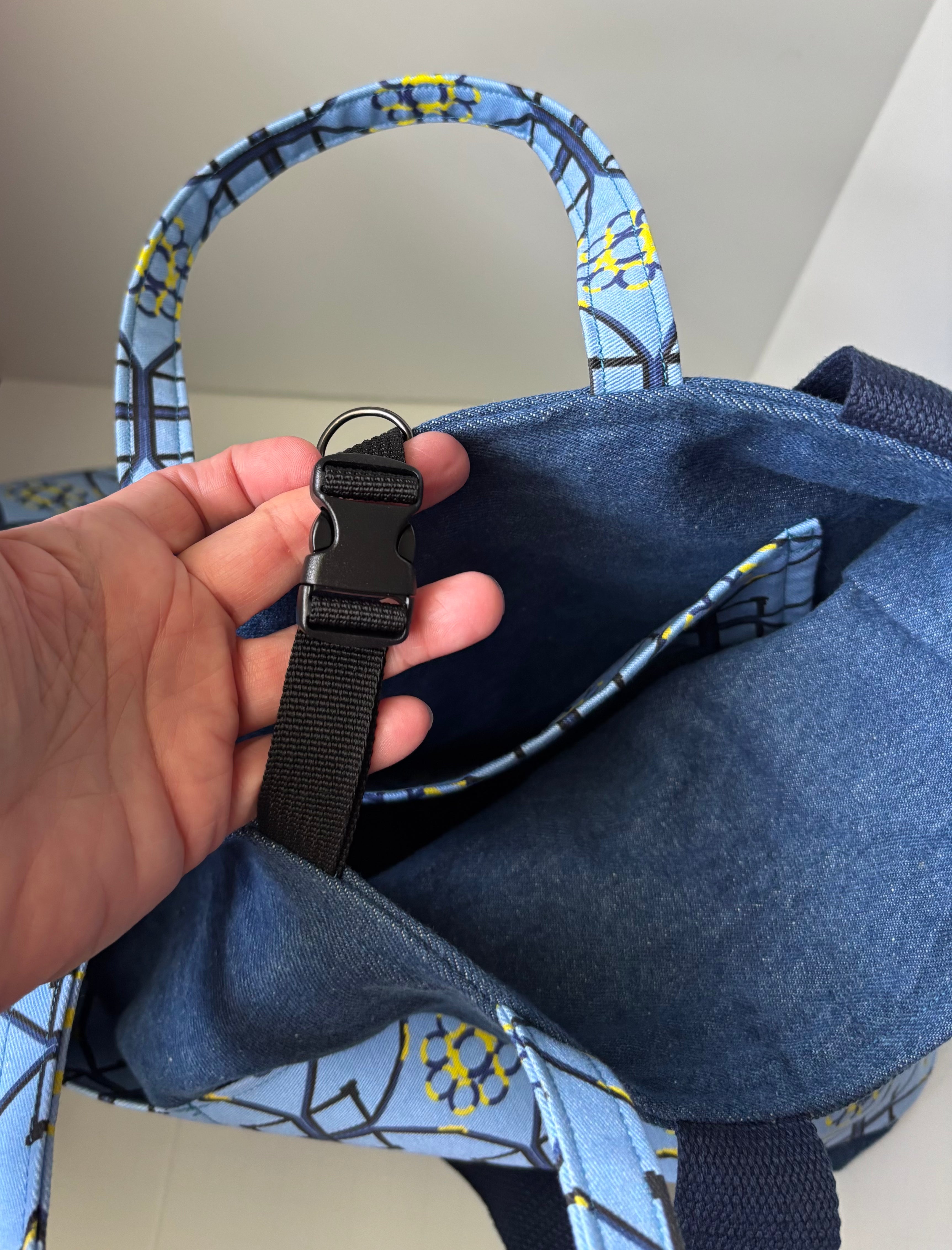 A close-up of a blue fabric tote bag with a black plastic buckle strap being held by a hand, showing the interior lining and patterned blue handles.