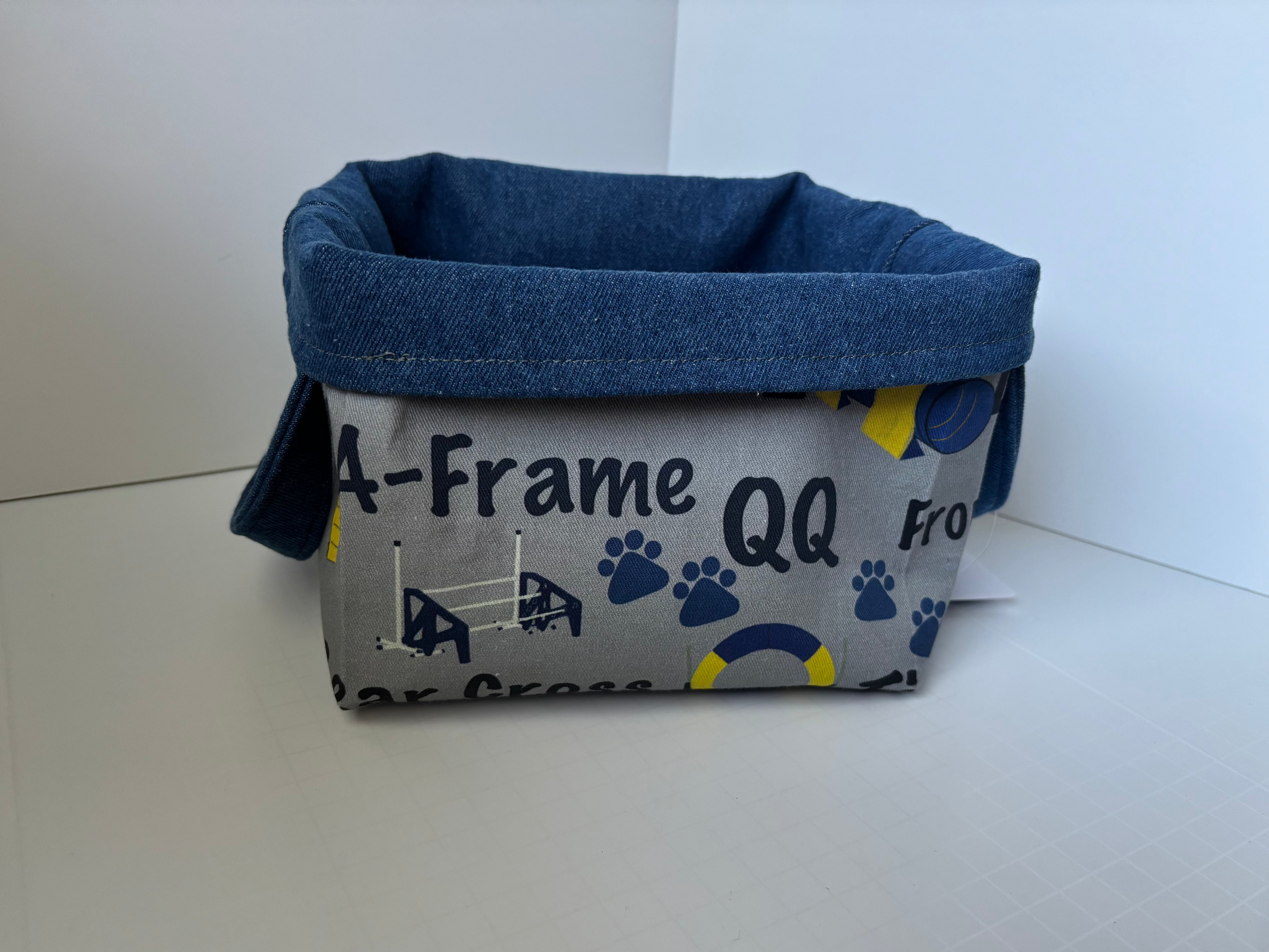 A fabric storage basket with a gray and navy blue patterned exterior featuring dog paw prints and text, and a solid navy blue fabric-lined interior.