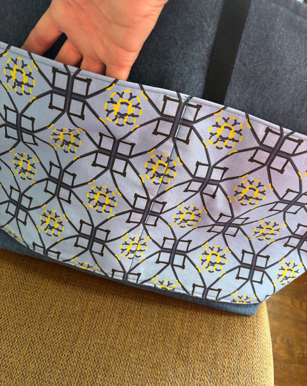 A close-up of a fabric tote bag with a geometric black and yellow pattern on a light blue background, held by a hand over a brown woven chair.