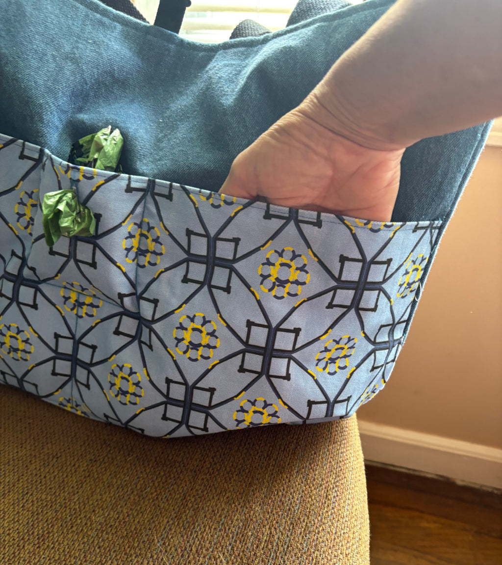 A close-up of a tote bag with a blue denim-like top section and a front pocket featuring a gray, black, and yellow geometric pattern. A hand is reaching into the bag, and the bag is resting on a textured tan chair in an indoor setting.