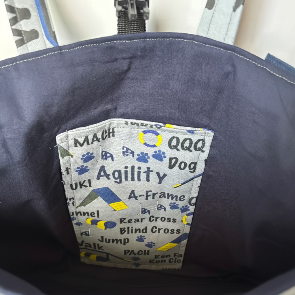 Close-up of the inside of a dark navy blue dog harness or bag, showing a printed white fabric tag with blue paw prints and text including 'MACH', 'Agility', 'Ooo Dog', and 'A-Frame'.