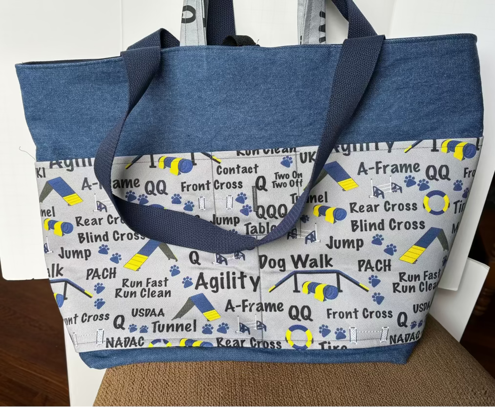 A fabric tote bag with a blue denim-like top and handles, and a light gray printed lower section featuring dog agility event terms and paw prints. The bag is photographed indoors against a white door.