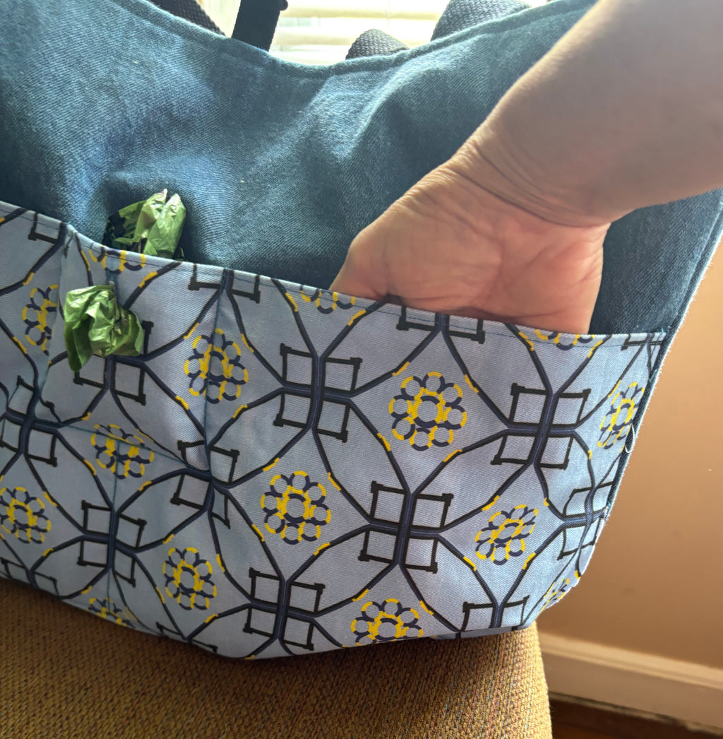 A close-up of a tote bag with a blue denim-like top section and a front pocket featuring a gray, black, and yellow geometric dog agility pattern. A hand is reaching into the bag, and the bag is resting on a textured tan chair in an indoor setting.