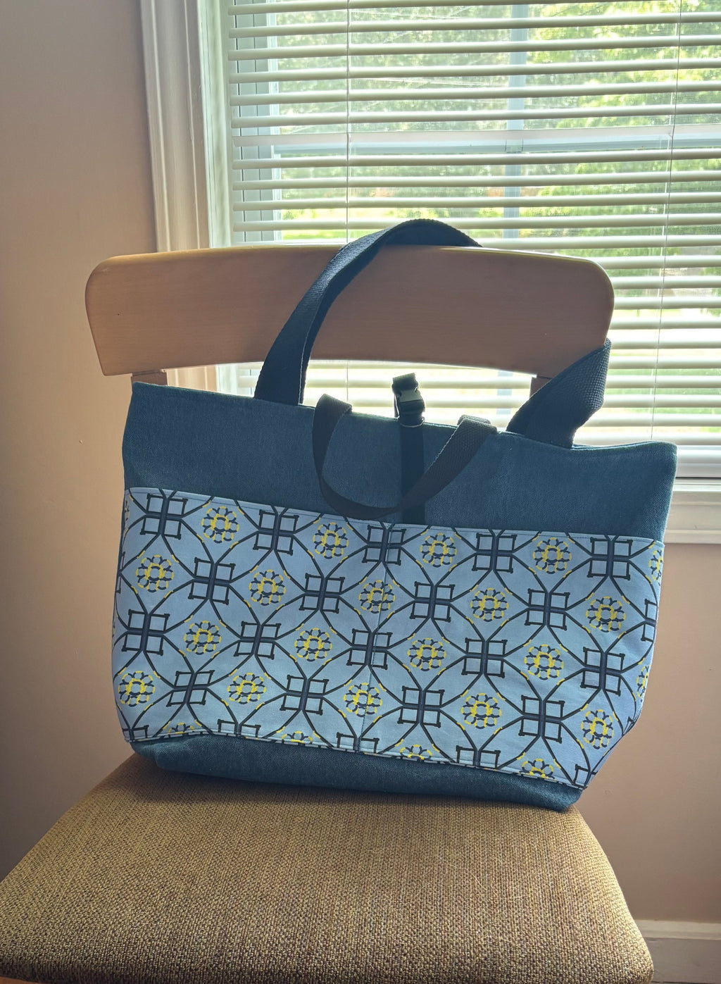A blue tote bag with a geometric dog agility patterned front panel sits on a wooden chair in front of a window with blinds.